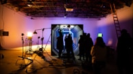 In a dimly lit studio, several silhouetted figures gather around an illuminated set piece resembling a futuristic corridor. Bright studio lights on stands cast shadows, and equipment like cameras and microphones are visible. The ceiling has an industrial look with exposed beams, and a ladder leans against the wall.