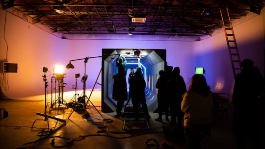 In a dimly lit studio, several silhouetted figures gather around an illuminated set piece resembling a futuristic corridor. Bright studio lights on stands cast shadows, and equipment like cameras and microphones are visible. The ceiling has an industrial look with exposed beams, and a ladder leans against the wall.
