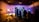 In a dimly lit studio, several silhouetted figures gather around an illuminated set piece resembling a futuristic corridor. Bright studio lights on stands cast shadows, and equipment like cameras and microphones are visible. The ceiling has an industrial look with exposed beams, and a ladder leans against the wall.