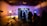 In a dimly lit studio, several silhouetted figures gather around an illuminated set piece resembling a futuristic corridor. Bright studio lights on stands cast shadows, and equipment like cameras and microphones are visible. The ceiling has an industrial look with exposed beams, and a ladder leans against the wall.