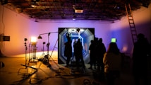 In a dimly lit studio, several silhouetted figures gather around an illuminated set piece resembling a futuristic corridor. Bright studio lights on stands cast shadows, and equipment like cameras and microphones are visible. The ceiling has an industrial look with exposed beams, and a ladder leans against the wall.