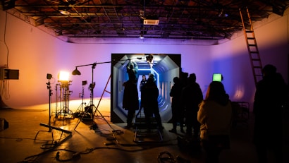 In a dimly lit studio, several silhouetted figures gather around an illuminated set piece resembling a futuristic corridor. Bright studio lights on stands cast shadows, and equipment like cameras and microphones are visible. The ceiling has an industrial look with exposed beams, and a ladder leans against the wall.