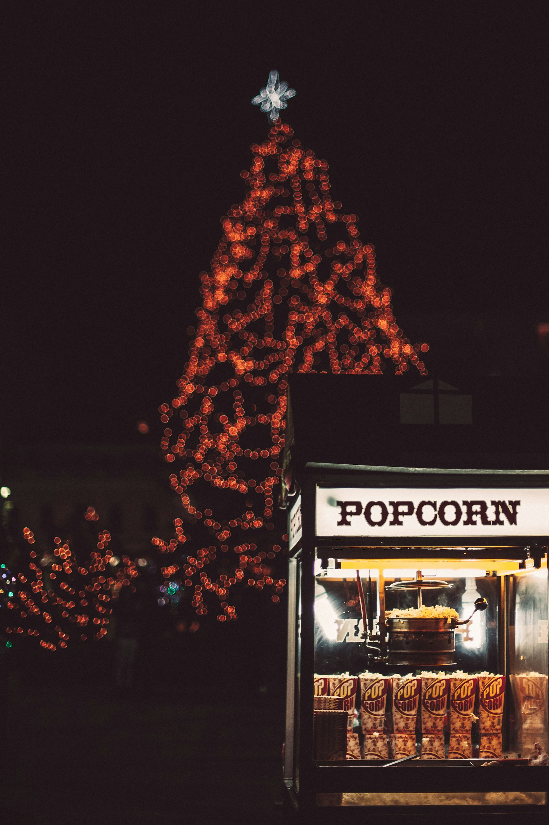 Bright festive lights arranged in the shape of a Christmas tree dominate the background, with a star on top. In the foreground, a popcorn machine is lit up, displaying bags of popcorn ready for sale. The scene captures a holiday ambiance, merging the warmth of a street vendor with seasonal decorations.