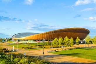 A modern, oval-shaped arena with a distinctive wooden exterior is set against a clear, sunny blue sky. Surrounding the structure is a neatly landscaped area with green grass, pathways, and a row of trees. The arena's unique architecture stands out prominently in a spacious, open environment.