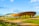 A modern, oval-shaped arena with a distinctive wooden exterior is set against a clear, sunny blue sky. Surrounding the structure is a neatly landscaped area with green grass, pathways, and a row of trees. The arena's unique architecture stands out prominently in a spacious, open environment.