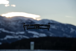 A sleek military drone flying over a rugged mountain landscape at sunset.