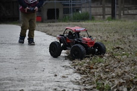 A child is playing with a red remote-controlled car on a paved path surrounded by grass and scattered leaves. The car has large wheels and a rugged design, suggesting it is suited for off-road use. The child's legs and hands are visible, indicating they are actively controlling the car.