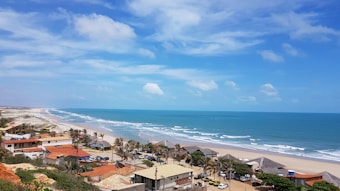 A picturesque coastal town with a beautiful sandy beach stretching along the shoreline. The image shows several buildings with red and beige roofs near the coastline, and the sea is a calm, deep blue with gentle waves. The sky is clear with some scattered white clouds, creating a serene and tranquil atmosphere. A few palm trees are visible around the buildings, contributing to the tropical vibe of the scene.