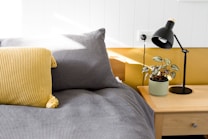 A cozy bedroom scene featuring a neatly made bed with a gray checkered duvet and a yellow knitted pillow. Beside the bed, there's a wooden nightstand supporting a black desk lamp and a potted plant with variegated leaves, adding a touch of nature to the setting. The room is well-lit with natural sunlight streaming in, highlighting the modern and minimalist design.