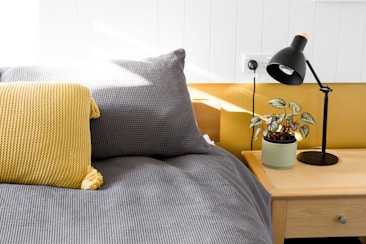 A cozy bedroom scene featuring a neatly made bed with a gray checkered duvet and a yellow knitted pillow. Beside the bed, there's a wooden nightstand supporting a black desk lamp and a potted plant with variegated leaves, adding a touch of nature to the setting. The room is well-lit with natural sunlight streaming in, highlighting the modern and minimalist design.