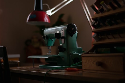 A vintage green sewing machine is positioned on a wooden table, surrounded by dim lighting. To the right, there is a wooden thread organizer filled with spools of various colors. Above the machine, a red lamp provides focused illumination. The overall setting suggests an intimate and quiet workspace dedicated to sewing.