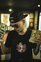 A person with long light-colored hair wearing a black cap and sunglasses holds a wad of cash in each hand. The t-shirt has a graphic design with the word 'explosive' on it. The background is blurred with dim lighting, giving the scene a moody atmosphere.