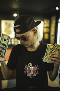 A person with long light-colored hair wearing a black cap and sunglasses holds a wad of cash in each hand. The t-shirt has a graphic design with the word 'explosive' on it. The background is blurred with dim lighting, giving the scene a moody atmosphere.