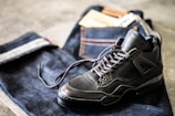 A pair of dark sneakers is placed on top of folded dark denim jeans with visible stitching and a leather patch. The scene conveys a casual and stylish aesthetic.