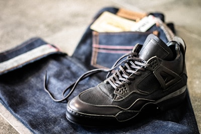 A pair of dark sneakers is placed on top of folded dark denim jeans with visible stitching and a leather patch. The scene conveys a casual and stylish aesthetic.