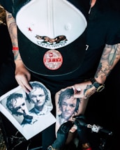 A person with tattooed arms is engaged in tattooing a realistic portrait on someone's leg. The scene includes printed images of the portrait being tattooed. The individual wears a baseball cap and has a tattoo machine in hand.
