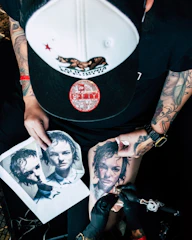 A person with tattooed arms is engaged in tattooing a realistic portrait on someone's leg. The scene includes printed images of the portrait being tattooed. The individual wears a baseball cap and has a tattoo machine in hand.