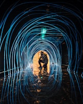 A person is squatting in the center of a tunnel-like structure made of glowing blue lines. The reflective floor mirrors the curved lines, creating an immersive visual experience. The tunnel leads towards a luminous area with warm lighting in the distance.