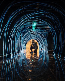 A person is squatting in the center of a tunnel-like structure made of glowing blue lines. The reflective floor mirrors the curved lines, creating an immersive visual experience. The tunnel leads towards a luminous area with warm lighting in the distance.