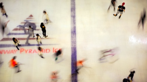 A group of people are ice skating on a rink, captured from above, resulting in a slightly blurred appearance due to movement. The ice surface displays a large logo or text near the center. Participants appear casually dressed, and the scene conveys a dynamic, bustling atmosphere.