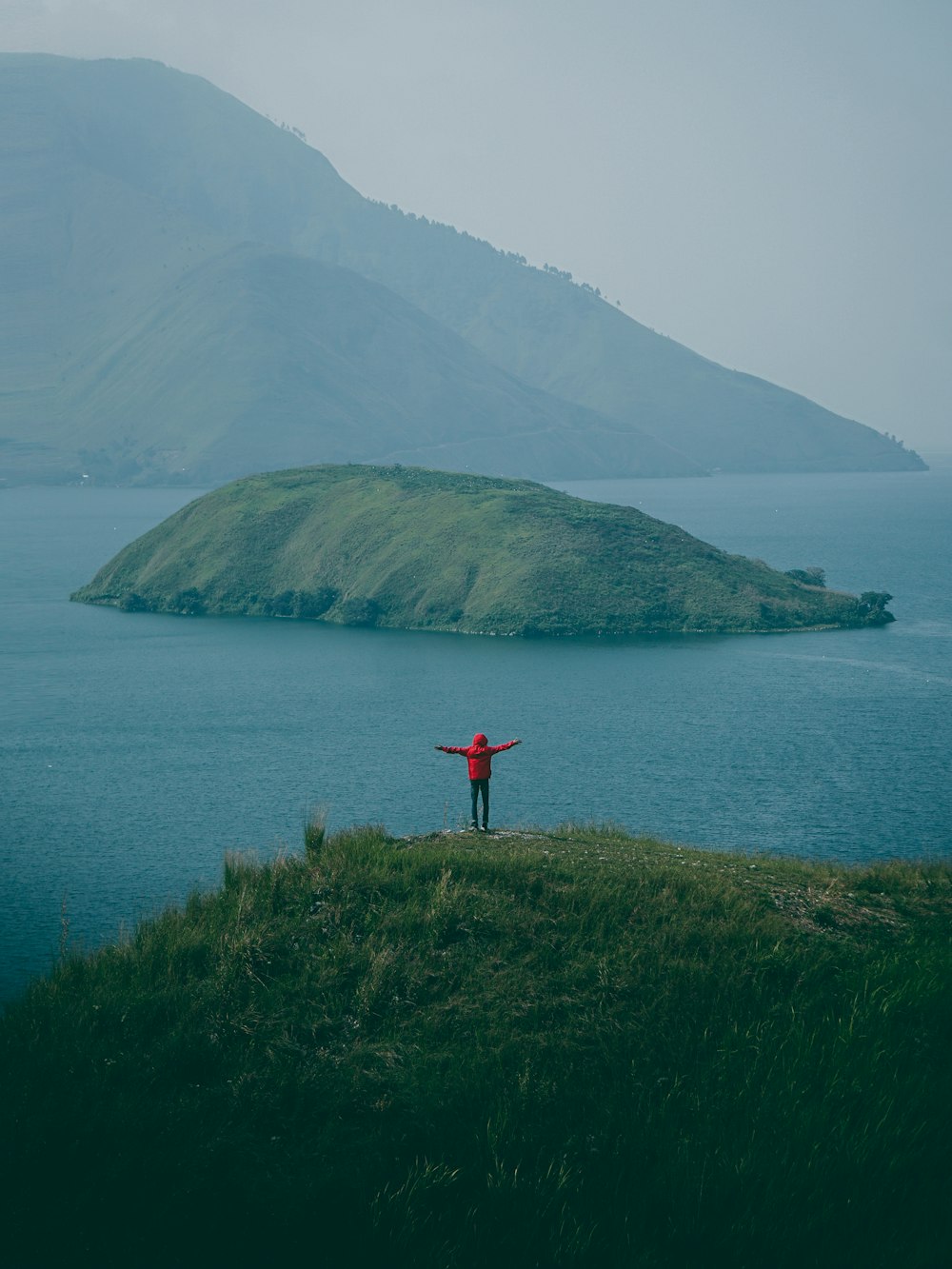 man in red jacket standing on hill facing island during daytime