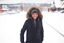 A smiling young person wrapped in a cozy winter jacket, standing against a snowy urban backdrop.