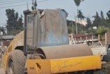 A large yellow construction vehicle with massive wheels and a cylindrical roller is stationed outdoors. The window of the vehicle’s cabin is slightly open, and the vehicle is partially covered with a draped, weathered tarp. In the background, utility poles, trees, and a blurred truck can be seen.