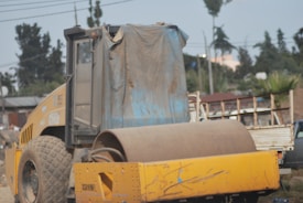 A large yellow construction vehicle with massive wheels and a cylindrical roller is stationed outdoors. The window of the vehicle’s cabin is slightly open, and the vehicle is partially covered with a draped, weathered tarp. In the background, utility poles, trees, and a blurred truck can be seen.