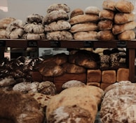 A variety of artisanal breads are neatly stacked on wooden shelves, featuring diverse textures and shapes. The assortment includes crusty round loaves, soft rolls, and rectangular loaves, some adorned with seeds.