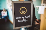 A chalkboard sign features a yellow smiling face above the text 'You're Up Next! Hang Tight.' It is positioned indoors, with a blurred figure seated in the background against a teal-colored wall.