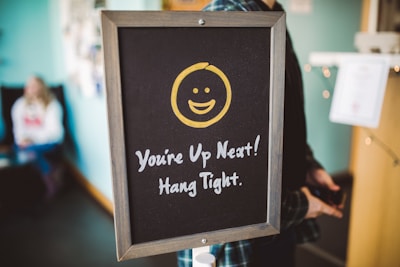 A chalkboard sign features a yellow smiling face above the text 'You're Up Next! Hang Tight.' It is positioned indoors, with a blurred figure seated in the background against a teal-colored wall.