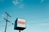 A cube-shaped sign with the word 'SURGERY' in red letters on a white background. Nearby, a tall utility pole and several power lines stretch across a bright blue sky with scattered clouds.