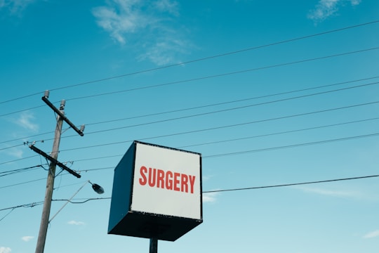 A cube-shaped sign with the word 'SURGERY' in red letters on a white background. Nearby, a tall utility pole and several power lines stretch across a bright blue sky with scattered clouds.