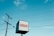 A cube-shaped sign with the word 'SURGERY' in red letters on a white background. Nearby, a tall utility pole and several power lines stretch across a bright blue sky with scattered clouds.