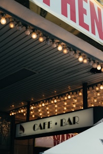 A marquee with bright round bulbs illuminates the entrance of a building. Below the lights, a sign reads 'Cafe • Bar,' suggesting a cozy or nightlife venue. Part of the tent or canopy edge appears at the bottom of the image.