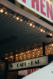 A marquee with bright round bulbs illuminates the entrance of a building. Below the lights, a sign reads 'Cafe &bull; Bar,' suggesting a cozy or nightlife venue. Part of the tent or canopy edge appears at the bottom of the image.