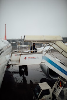 A person is standing on the jet bridge connected to a China Eastern airplane, with a wet tarmac visible below. The airplane and terminal are in the background.