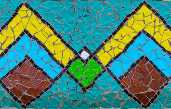 A colorful mosaic composed of various geometric shapes. The design includes large diamond patterns with vibrant shades of turquoise, yellow, blue, green, brown, and small sections of white. The mosaic pieces are irregularly shaped and fitted closely together to form a cohesive pattern.