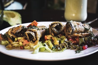 Three rolled, crispy-fried vegetable wraps placed on a bed of mixed salad greens and diced vegetables. The wraps are garnished with herbs, and the plate is placed on a dimly-lit dining table. In the background, there is an avocado half and a creamy drink in a tall glass.