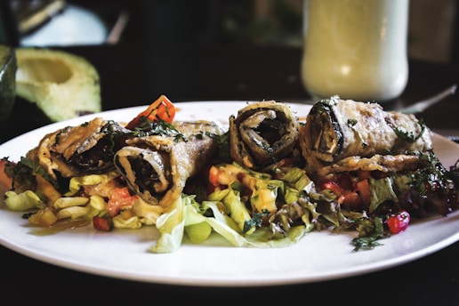 Three rolled, crispy-fried vegetable wraps placed on a bed of mixed salad greens and diced vegetables. The wraps are garnished with herbs, and the plate is placed on a dimly-lit dining table. In the background, there is an avocado half and a creamy drink in a tall glass.