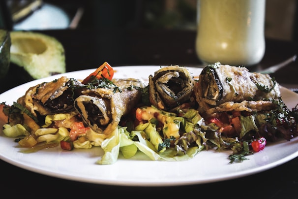 Three rolled, crispy-fried vegetable wraps placed on a bed of mixed salad greens and diced vegetables. The wraps are garnished with herbs, and the plate is placed on a dimly-lit dining table. In the background, there is an avocado half and a creamy drink in a tall glass.