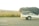 A white trailer with a black net covering its contents travels on a highway. The landscape shows grassy fields and distant houses in the background under an overcast sky.