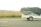 A white trailer with a black net covering its contents travels on a highway. The landscape shows grassy fields and distant houses in the background under an overcast sky.