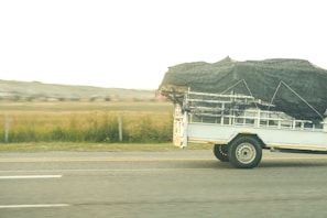 A white trailer with a black net covering its contents travels on a highway. The landscape shows grassy fields and distant houses in the background under an overcast sky.