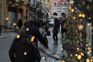 A bustling European cobblestone street with people walking and shopping. Twinkling lights decorate the trees lining the path, creating a festive atmosphere. Buildings with balconies and shop windows frame the scene, adding a cozy urban charm.