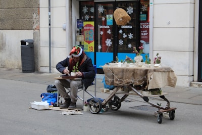 A person wearing a festive hat and a garland is sitting on a portable chair on the sidewalk, engaging in some activity on a clipboard. In front of them is a makeshift table on wheels, covered with a brown cloth and decorated with small plant arrangements, possibly cacti. The scene is set outside a building with glass doors adorned with snowflake decals.
