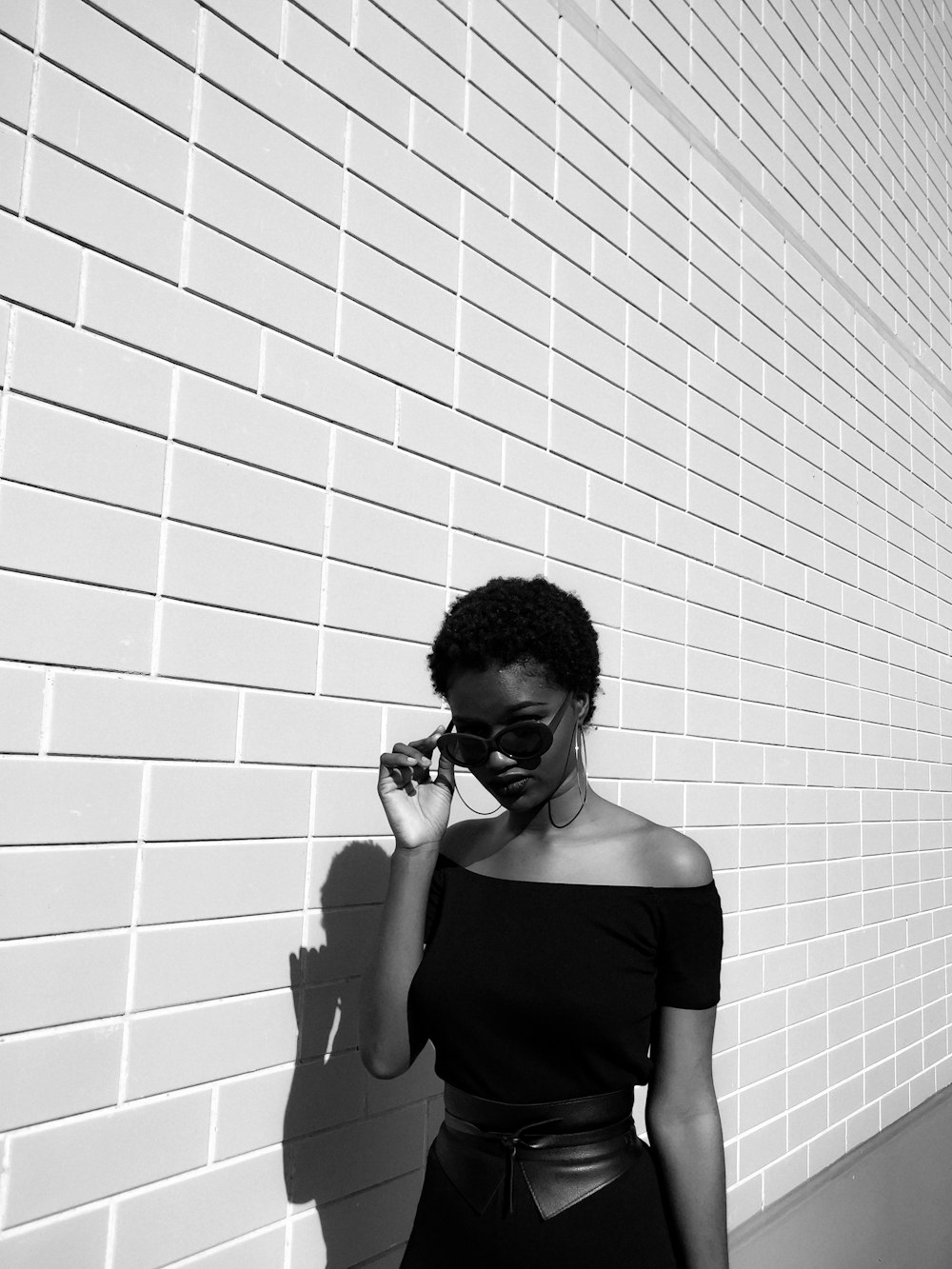 grayscale photography of woman wearing off-shoulder dress and sunglasses next to brick wall