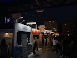 A nighttime street festival with multiple booths and bright string lights illuminating the area. There are people gathered in front of brightly lit stalls, and signs written in a non-English script are visible. The ambiance is lively and bustling with various decorative elements hanging above.
