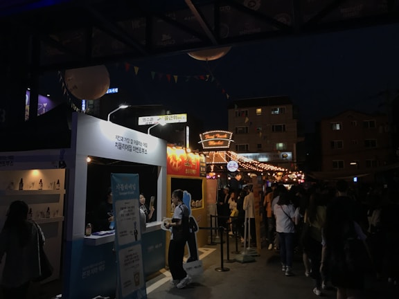 A nighttime street festival with multiple booths and bright string lights illuminating the area. There are people gathered in front of brightly lit stalls, and signs written in a non-English script are visible. The ambiance is lively and bustling with various decorative elements hanging above.
