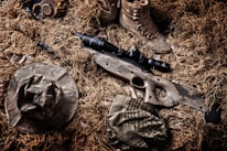 A camouflaged sniper rifle lies amidst a backdrop of tactical gear and a ghillie suit. Surrounding the rifle are a pair of tan combat boots, a camouflage hat, and other military accessories. The colors and textures blend with the natural environment, indicating a setup for concealment in an outdoor setting.